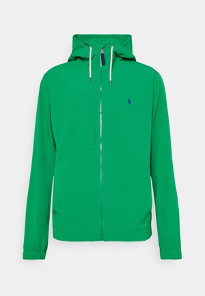 Veste à capuche verte avec fermeture éclair sur le devant, cordons blancs, poignets élastiques et petit logo sombre sur le côté gauche de la poitrine.
