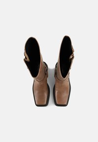 Bottes hautes en cuir marron avec bouts carrés, semelles noires et brides décoratives, vues de dessus sur un fond blanc.