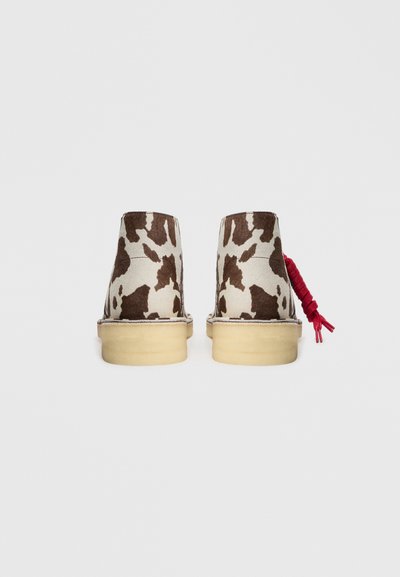 Brązowo-białe zamszowe botki w wzór przypominający skórę krowy, z naturalną gumową podeszwą i czerwoną ozdobną kokardą z tyłu.