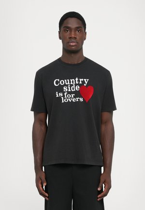 Young Black man standing, wearing black t-shirt with white text "Countryside is for lovers" and red heart graphic on front.