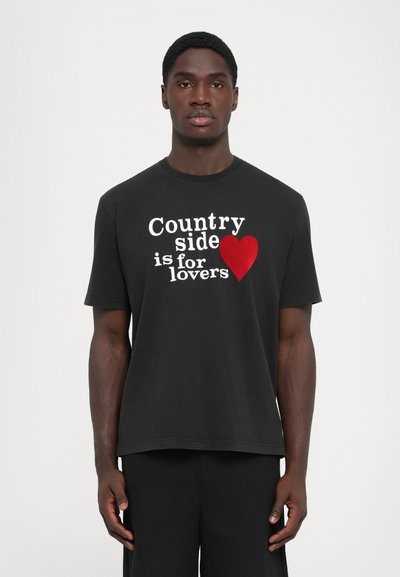 Młody czarnoskóry mężczyzna stoi, ubrany w czarną koszulkę z białym napisem "Countryside is for lovers" oraz graficznym czerwonym sercem z przodu.