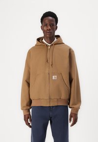 Haut à capuche zippé marron en mélange de coton, avec une poche avant et des cordons sur la capuche. Porté sur une chemise blanche à col.