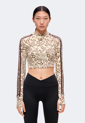 Jeune femme aux cheveux noirs et raides porte un crop top à manches longues imprimé léopard et des leggings noirs taille haute, debout devant un fond uni.