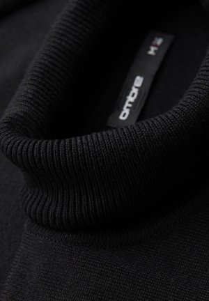 Zwarte trui met een hoge, geribbelde kraag. Het materiaal lijkt textuur te hebben, met een gladde body. Het binnenlabel toont de merknaam en maat.