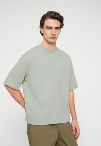 Νεαρός άνδρας με καστανά κυματιστά μαλλιά, που φοράει άνετη ανοιχτοπράσινη κοντομάνικη μπλούζα και χακί παντελόνι, στέκεται μπροστά σε απλό λευκό φόντο.