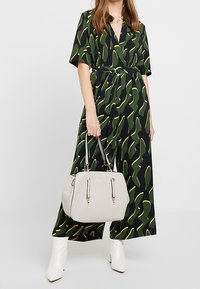 Nainen, jolla on vihreä, musta ja keltainen kuvioitu vyötäröltä kiristettävä mekko, pitää käsissään beigeä käsilaukkua ja hänellä on valkoiset nilkkasaappaat. Hän seisoo yksinkertaista taustaa vasten.