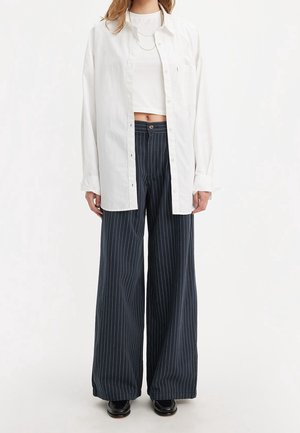 Mujer con camisa blanca holgada abotonada sobre un crop top blanco, pantalones anchos de rayas azul marino y zapatos negros, de pie sobre un fondo blanco.