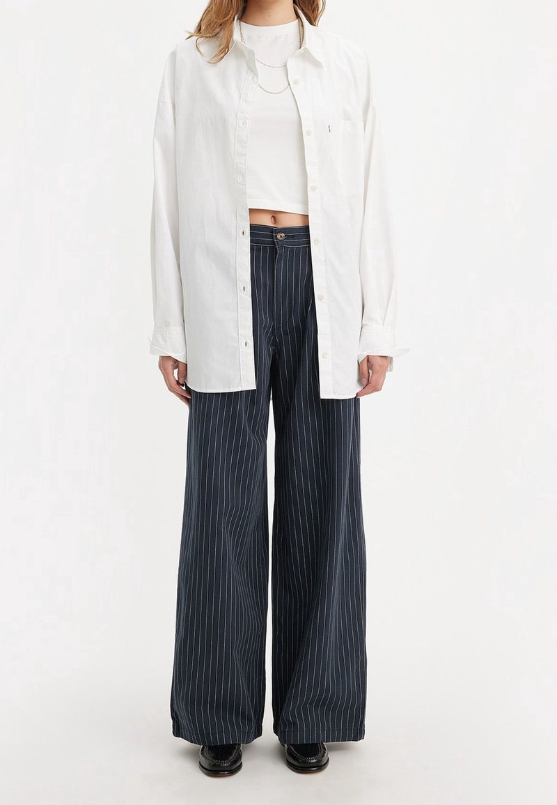Femme portant une chemise blanche ample boutonnée par-dessus un crop top blanc, pantalon large à rayures bleu marine et chaussures noires, debout sur fond blanc.