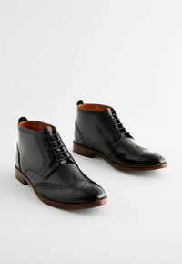 Botas de cuero negras hasta el tobillo con puntera perforada y diseños laterales, suela de cuero marrón y forro interior naranja en contraste.