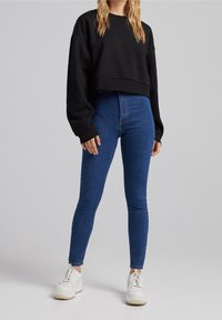 Svart croppad sweatshirt med sänka axlar, matchad med högmidjade blå skinny jeans och vita kraftiga sneakers. Slät textur och avslappnad design.