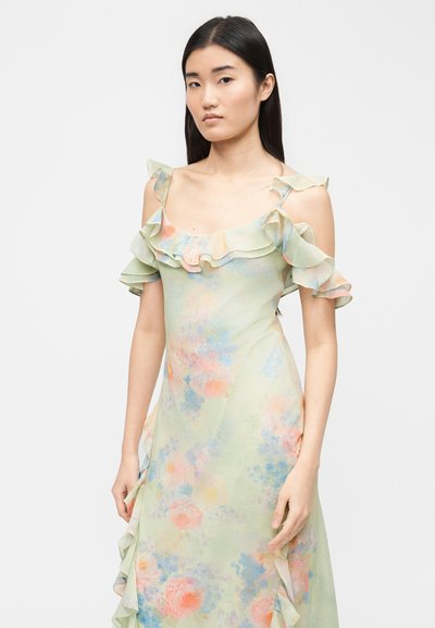 Νεαρή γυναίκα με μακριά σκούρα μαλλιά, ντυμένη με παστέλ πράσινο λουλουδάτο φόρεμα με βολάν στους ώμους που αφήνουν ακάλυπτους τους ώμους, στέκεται μπροστά από ένα απλό φόντο.