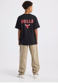 Camiseta negra de manga corta con el texto "BULLS" en rojo y el logo en la parte trasera; combinada con pantalones beige claro de corte relajado y zapatillas blancas.