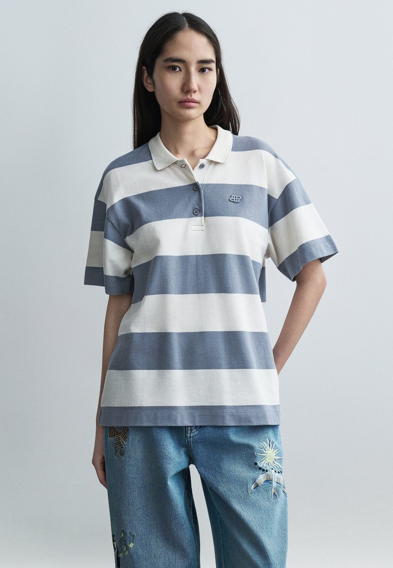 Polo rayé en mélange de coton doux, présentant des rayures horizontales bleues et crème, un col et trois boutons. Design décontracté et confortable.