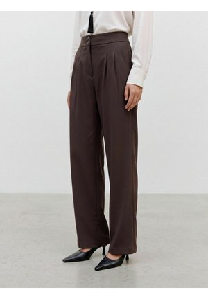 Femme portant un pantalon large taille haute marron foncé avec des plis et des talons noirs pointus, debout sur un sol gris.
