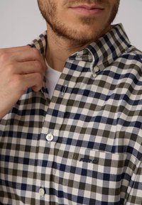 Chemise à carreaux avec col boutonné, présentant des carrés bleus marine, verts et blancs, avec une poche poitrine et un logo brodé subtil. Texture en coton lisse.