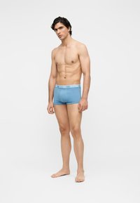 Άνδρας χωρίς πουκάμισο, όρθιος ξυπόλητος με ανοιχτο μπλε μπόξερ με λευκή ζώνη μέσης με τη μάρκα "MOSCHINO" σε απλό λευκό φόντο.