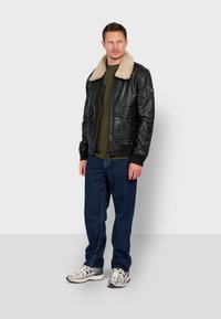 Černá kožená bomber jacket s béžovým beránkovým límcem, dvěma předními kapsami, nošená přes zelenou košili a modré džíny, doplněna bílými teniskami.