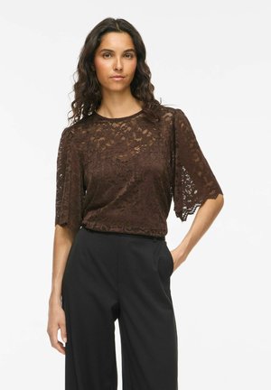 Femme portant un haut en dentelle à manches courtes marron foncé avec des motifs floraux et un pantalon noir taille haute, debout avec une main dans la poche.