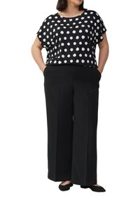 Vrouw in een zwart top met korte mouwen met stippen, zwarte wijde broek en zwarte puntige schoenen met haar handen in de zakken.