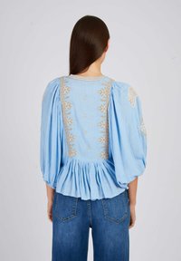 Lichtblauwe blouse met pofmouwen, geborduurde details in beige, geplooide achterkant en een ronde halslijn. Gecombineerd met blauwe denim jeans.