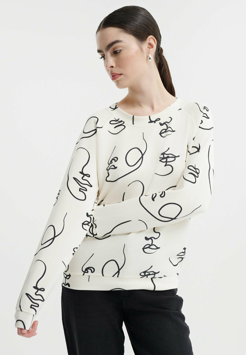 Sudadera blanca con dibujos de caras abstractas en línea negra, escote redondo y mangas largas, combinada con pantalones negros.