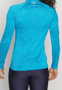Modrý sportovní top s dlouhým rukávem vyrobený z texturované tkaniny odvádějící vlhkost. Má vysoký límec a přiléhavý střih s raglánovými rukávy.