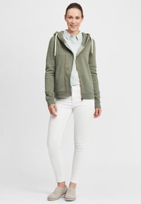 Green zip-up hoodie with drawstrings, side pockets, and brown accents, worn over a light blue striped shirt and white pants.