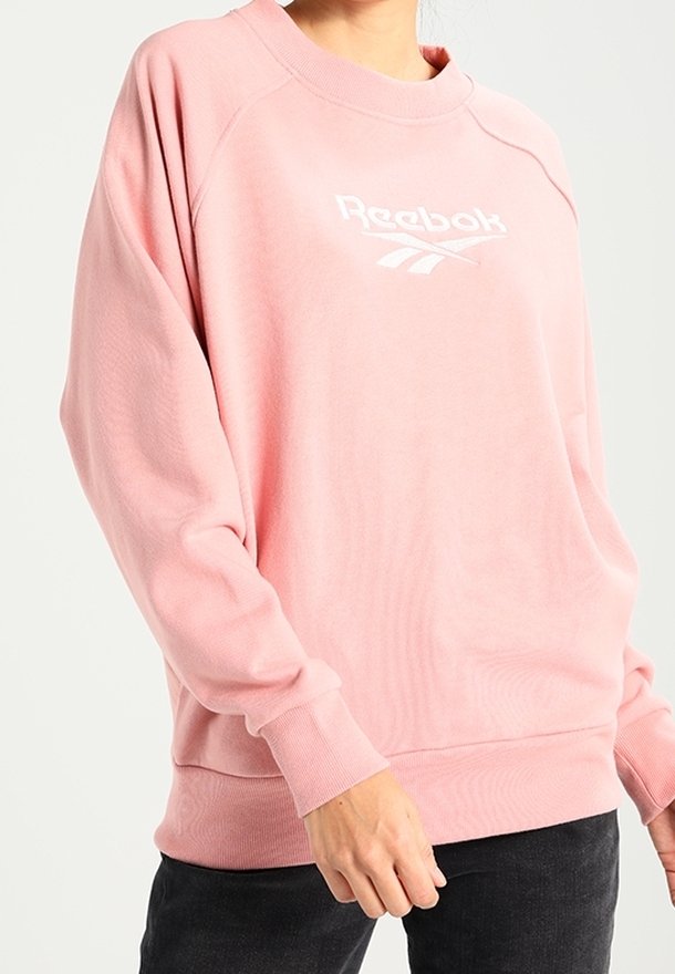Personne portant un sweatshirt ample rose clair Reebok avec logo blanc, assorti d’un pantalon noir, debout devant un fond clair uni.