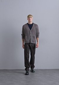 Grå ruggad cardigan med fickor över en svart skjorta, i kombination med mörka byxor och svarta skor. Modellen står mot en neutral bakgrund.
