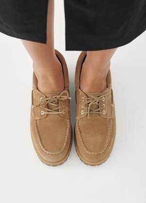 Brązowe zamszowe buty z okrągłym noskiem, wyposażone w teksturowane sznurowadła i złocone przelotki, na jasnym tle.