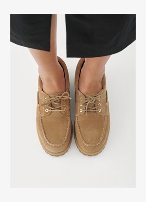 Pantofi din piele întoarsă maro cu vârf rotund, cu șireturi texturate și orificii aurii, așezați pe un fundal deschis.