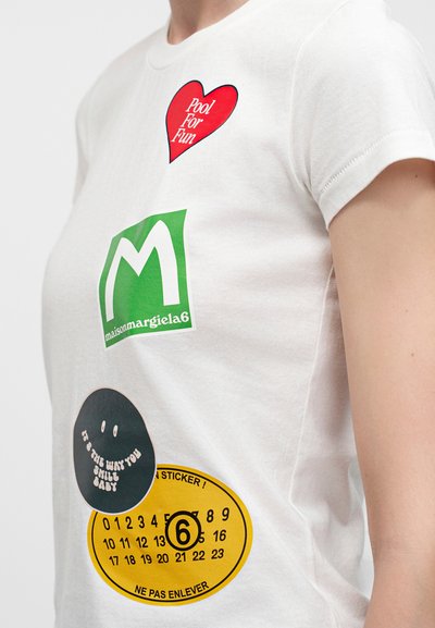 Λευκό μπλουζάκι με κόκκινη καρδιά που γράφει "Pool For Fun", πράσινο λογότυπο Maison Margiela, μαύρο χαμογελαστό πρόσωπο και κίτρινη κυκλική αυτοκόλλητη ετικέτα με αριθμούς και κείμενο.