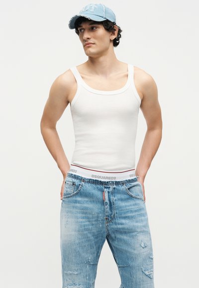 Νέος άνδρας που φοράει ανοιχτό μπλε καπέλο, λευκή φανέλα και ξεβαμμένο μπλε τζιν με τα χέρια στις τσέπες, κοιτάζοντας προς τα πλάγια σε απλό φόντο.