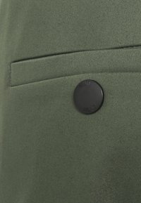 Zwarte ronde knop naast een horizontale gleufzak op een olijfgroen gestructureerd stof, waarschijnlijk onderdeel van een kledingstuk.