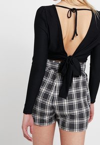 Schwarzes langärmliges Oberteil mit tiefem V-Ausschnitt auf der Rückseite und Bindeschluss, kombiniert mit hochgeschnittenen karierte Shorts in Schwarz und Weiß.