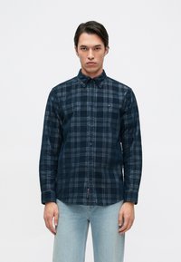 Camisa de cuadros azul marino con mangas largas, cuello abotonado y un logo en el pecho. Presenta una tela texturada y un diseño de corte recto.
