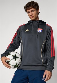 Mørkegrå hoodie med røde og guld accenter, der har en justerbar hætte, guldstriber og Olympique Lyonnais-logoet. Spiller holder en fodbold.