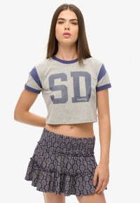 Graues, kurz geschnittenes T-Shirt mit blauen Ärmeln, versehen mit fetten "SD"-Buchstaben. Kombiniert mit einem dunklen, gemusterten ausgestellten Rock mit Rüschen.