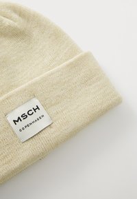 Strikket hue i lys beige med en blød tekstur. Har et mærke med "MSCH COPENHAGEN" i sort, synligt på den foldede kant.