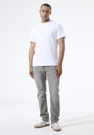 Weißes Baumwoll-T-Shirt mit kurzen Ärmeln, kombiniert mit grauer Denim-Jeans und grauen Sneakern mit weißen Akzenten, stehend vor einem schlichten Hintergrund.