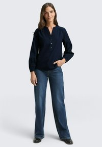 Blusa a maniche lunghe blu navy con scollo a V e tre bottoni, abbinata a jeans blu a gamba larga. La blusa presenta una leggera arricciatura alle spalle.