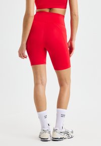Persona vestida con pantalones cortos deportivos de cintura alta de color rojo brillante, calcetines blancos de marca y zapatillas de correr blancas, de pie contra un fondo blanco liso.
