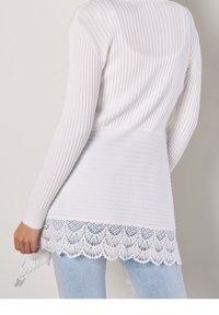 Žena v bílém žebrovaném svetr s krajkovým lemem a světle modrých džínách, stojící na jednobarevném světlém pozadí.