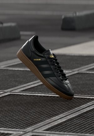 Sort Adidas Spezial sneaker med gummisål, der svæver over metalrist på urban fortov.