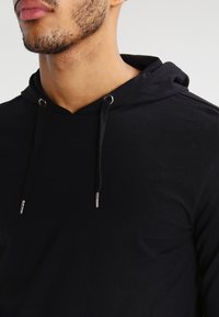 Černá bavlněná mikina s kapucí, vybavená šňůrkou se stříbrnými kovovými očky, příjemnou texturou a slim fit střihem, který zakrývá krk a ramena.