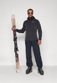 L'ensemble de ski se compose d'une veste noire avec des accents orange, de pantalons bleu marine et de bottes de ski noires ; il tient des skis marron avec des fixations.