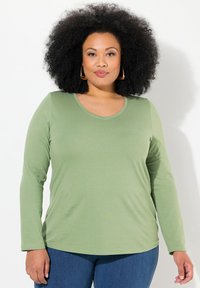 Dlouhý rukáv, top s V-výstřihem ve světle zelené látce s hladkým povrchem. V kombinaci s tmavě modrými džíny s přiléhavým střihem.