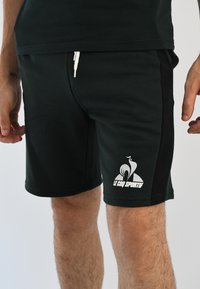 Shorts verde oscuro de mezcla de algodón, con un panel lateral negro en contraste y un logo blanco en la pierna izquierda. Cintura elástica con cordón.