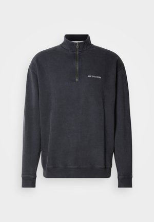 Grijs katoenmix pullover met rits tot kwart. Heeft een geribde kraag en manchetten, met een klein wit logo op de borst. Glad textuur.