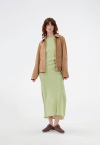 Καφέ σουέτ μπουφάν με γιακά και μπροστινές τσέπες, συνδυασμένο με ένα μακρύ, ανοιχτό πράσινο σατέν φόρεμα και σκούρα καφέ παπούτσια.